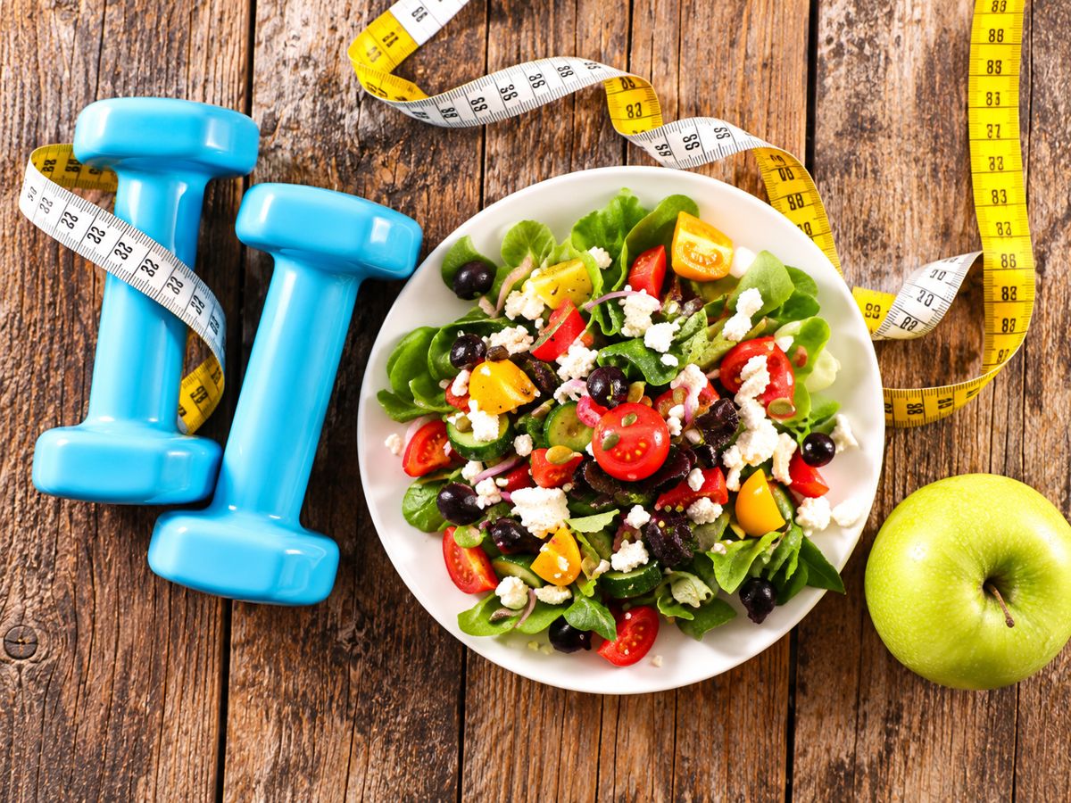 Dumbbells, a fresh salad, and an apple beside a measuring tape